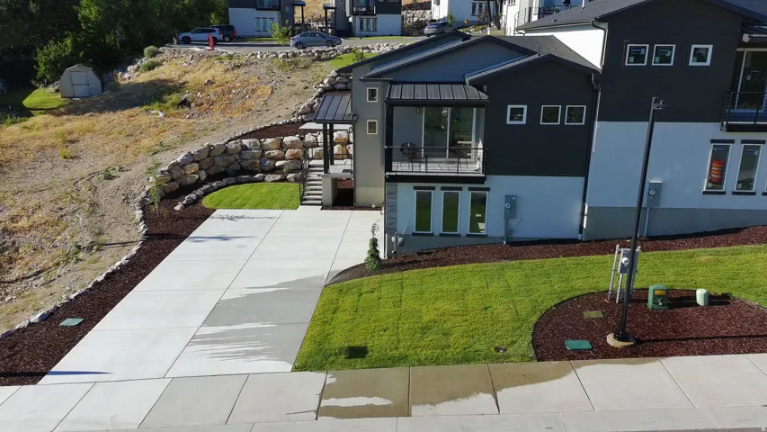 Contemporary exterior featuring dark and light siding, a spacious concrete driveway, manicured lawns, a stone retaining wall, and a covered balcony