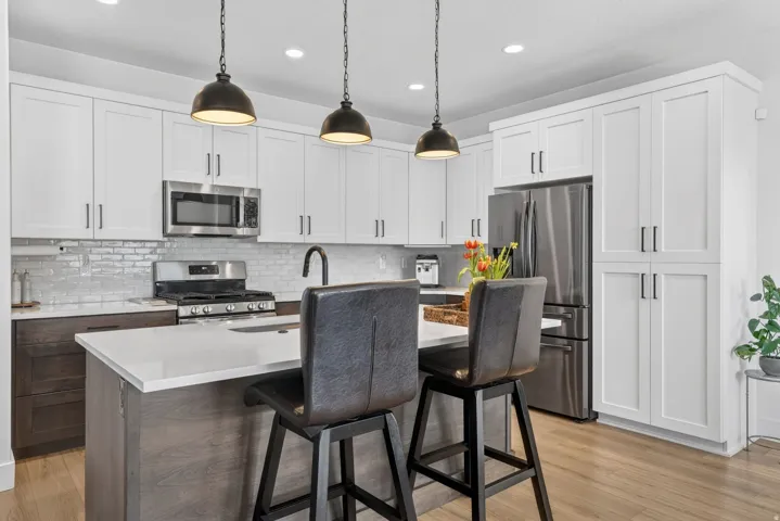 Two tone kitchen with stainless steel appliances, a kitchen island with sink, a kitchen bar, decorative backsplash, and decorative light fixtures