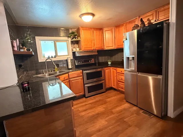 Kitchen featuring wood-finish flooring, wood cabinetry, dark countertops, a stainless steel double oven, and a stainless steel refrigerator