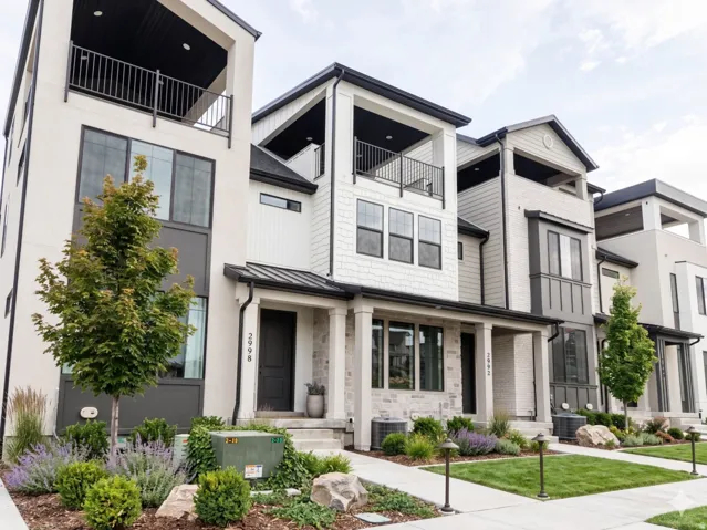 Modern townhome exterior featuring an open-air balcony, a covered porch entry, stacked stone accents, dark frame windows, and manicured landscaping