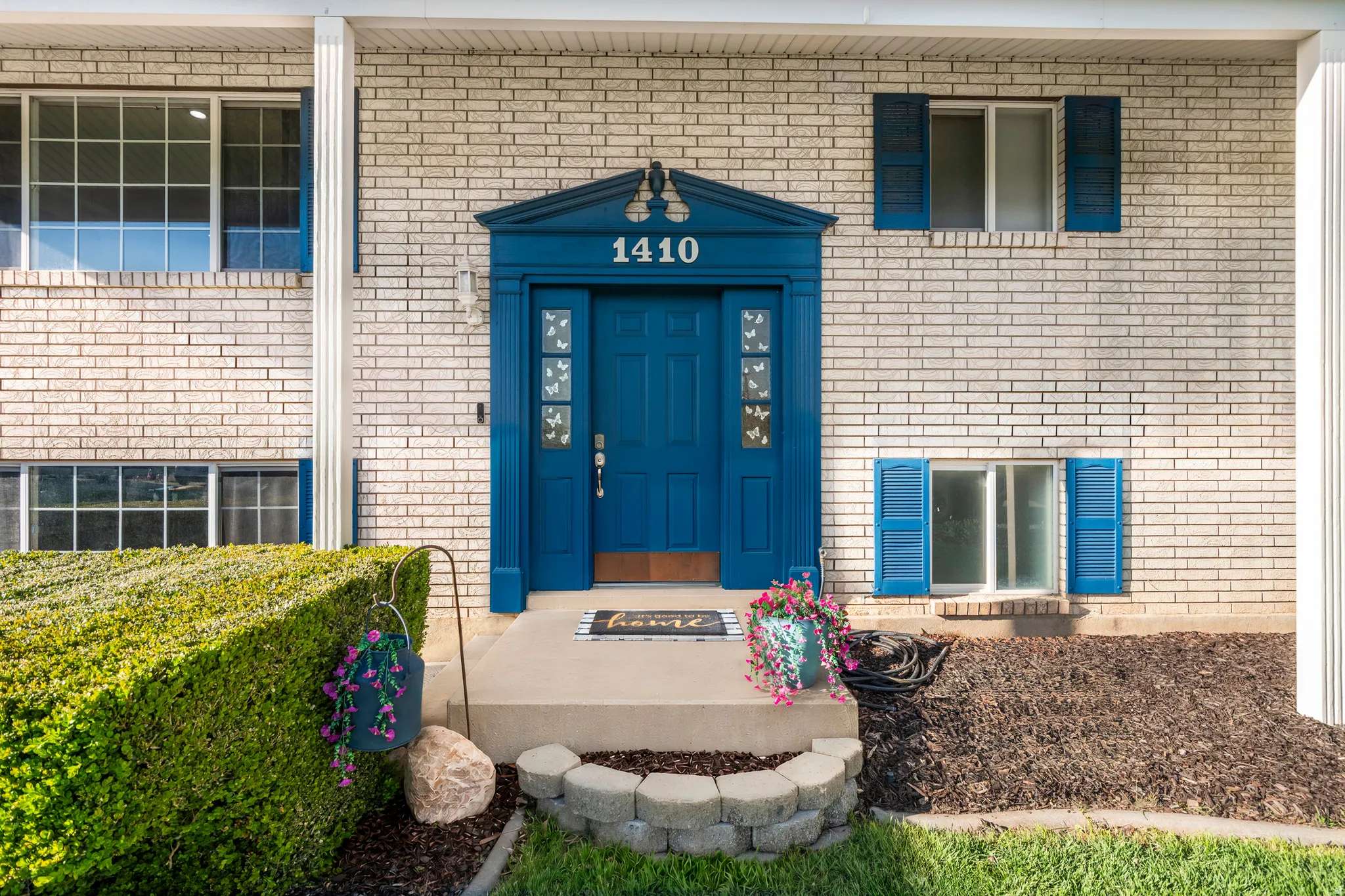 Light-colored brick exterior featuring a vibrant blue front door with decorative trim, complementary blue window shutters, and a concrete front porch