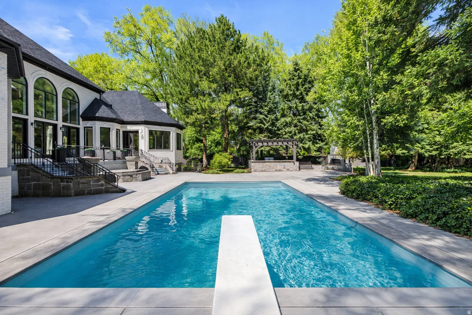 Saline swimming pool with a patio area, a diving board, view of wooded area, and a pergola with outdoor kitchen.