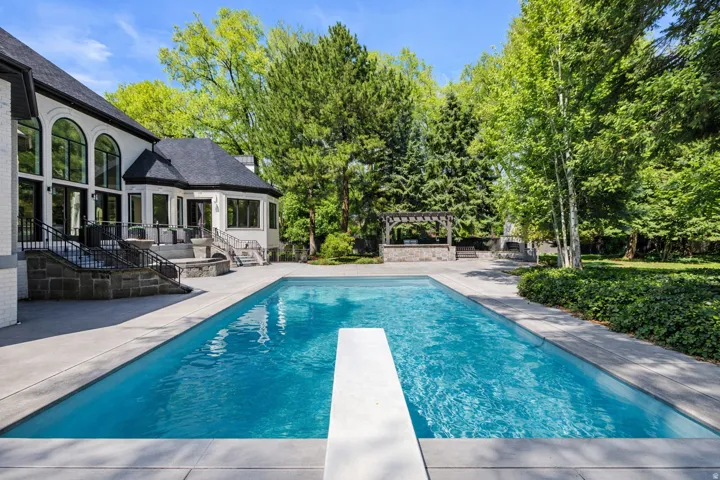Saline swimming pool with a patio area, a diving board, view of wooded area, and a pergola with outdoor kitchen.