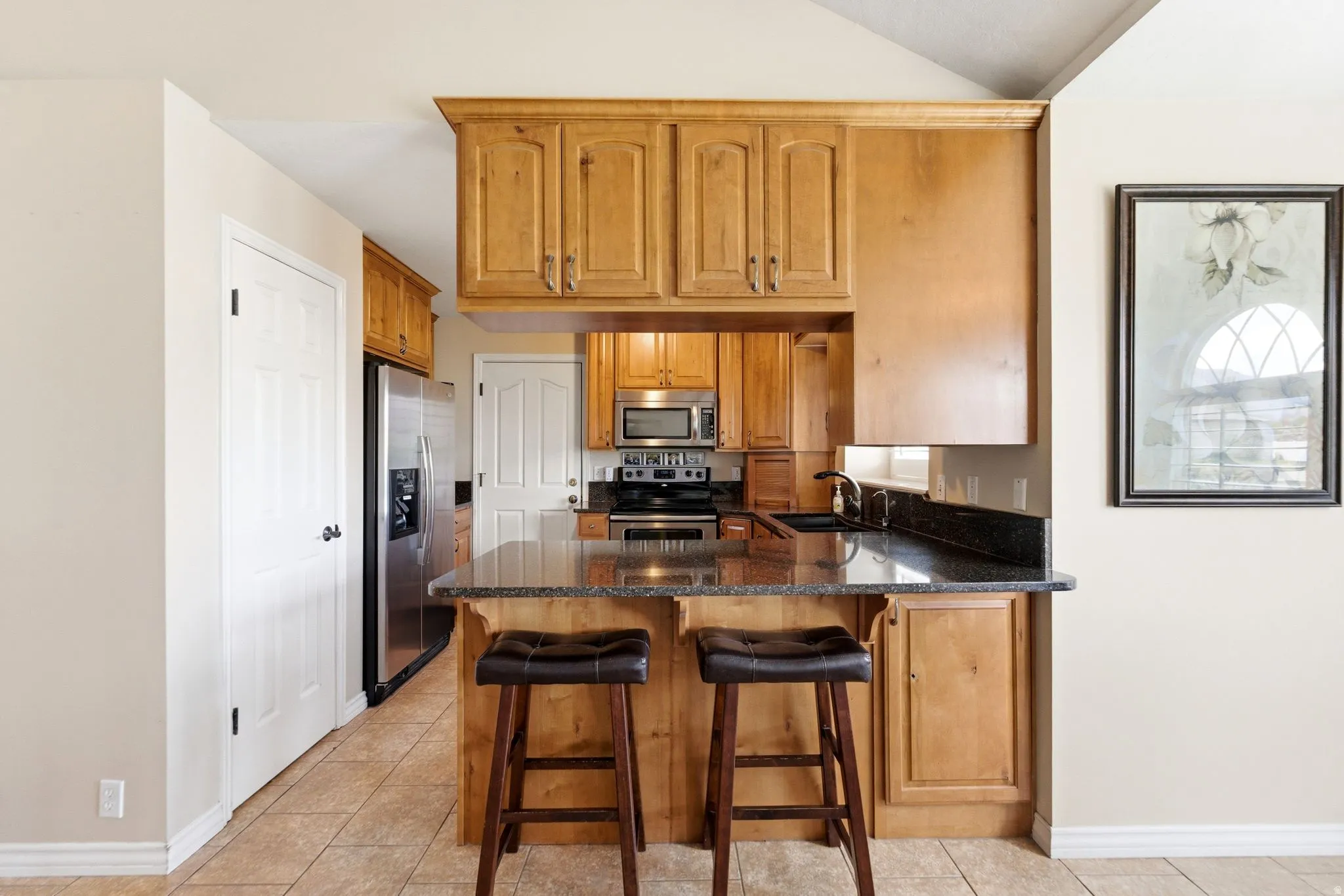 Kitchen with dark stone counters, a peninsula, stainless steel appliances, a kitchen bar, and wood finish cabinets