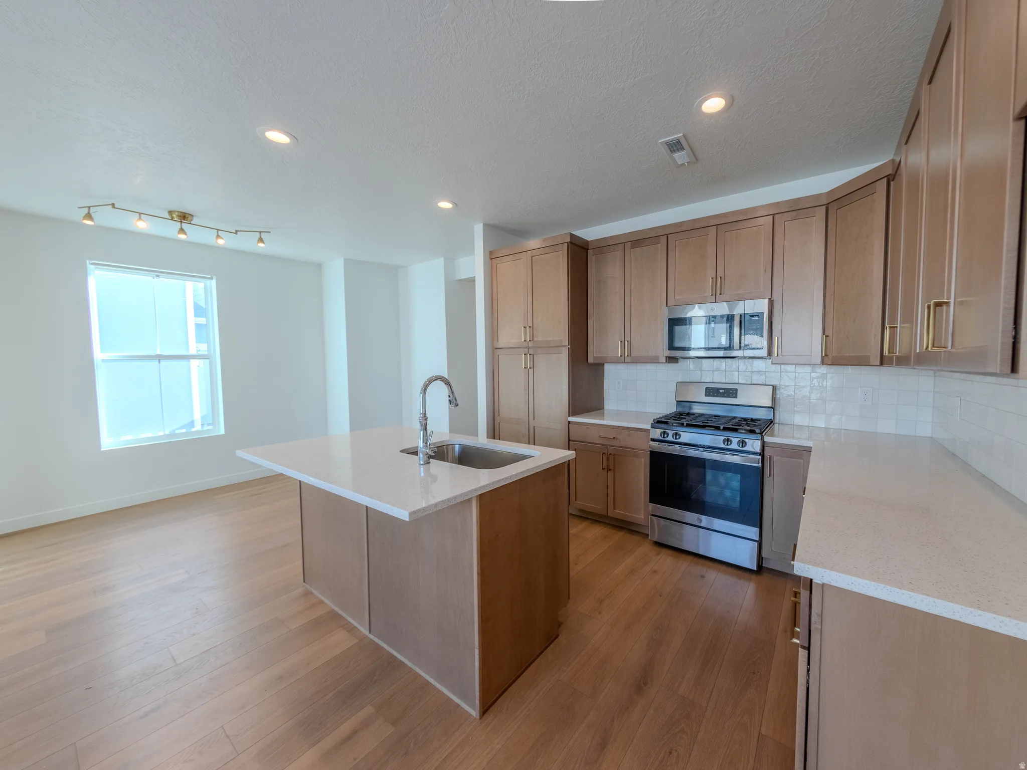 Kitchen with stainless steel appliances, a center island with sink, light stone counters, decorative backsplash, and light wood-style flooring