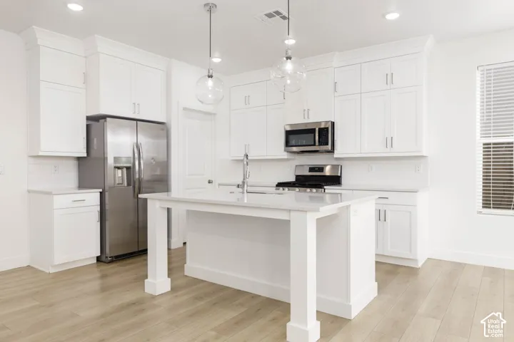 Kitchen featuring white cabinetry, stainless steel appliances, a center island with a sink, pendant lighting, and wood-finish flooring