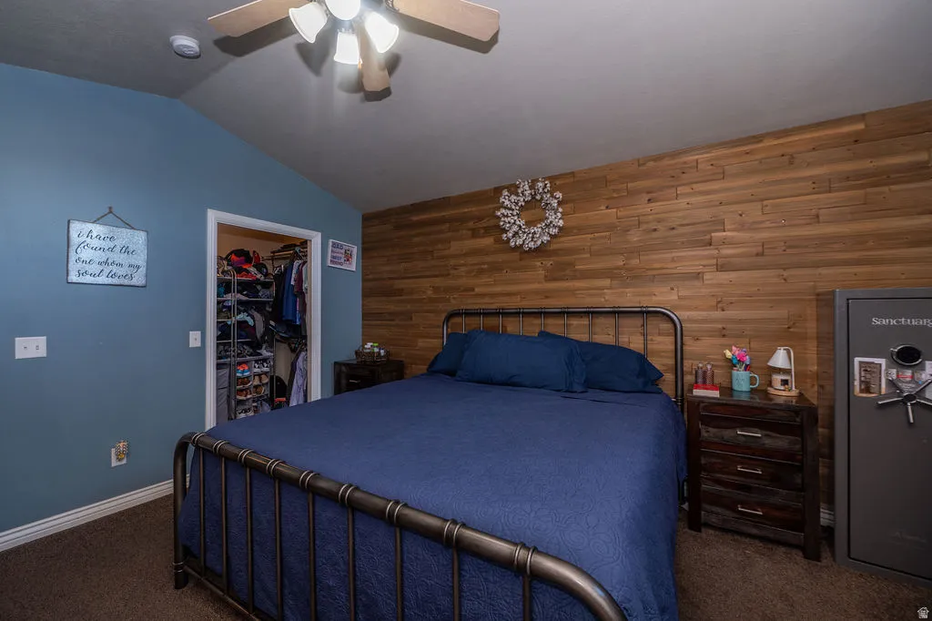 Bedroom featuring wood walls, a spacious closet, dark carpet, vaulted ceiling, and ceiling fan