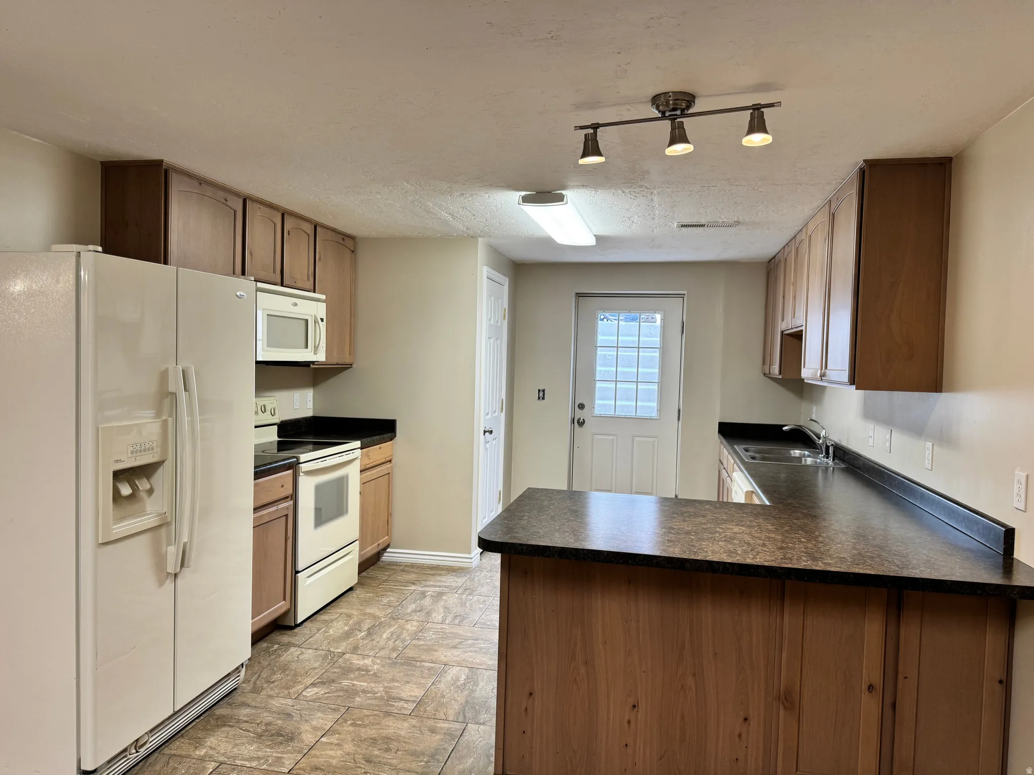 Kitchen with white appliances, a peninsula, dark countertops, a textured ceiling, and stone finish flooring