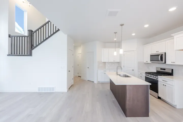 Open-concept design featuring wood-finish flooring, a kitchen island with pendant lighting, white cabinetry, stainless steel appliances, and a staircase with dark wood-finish railings