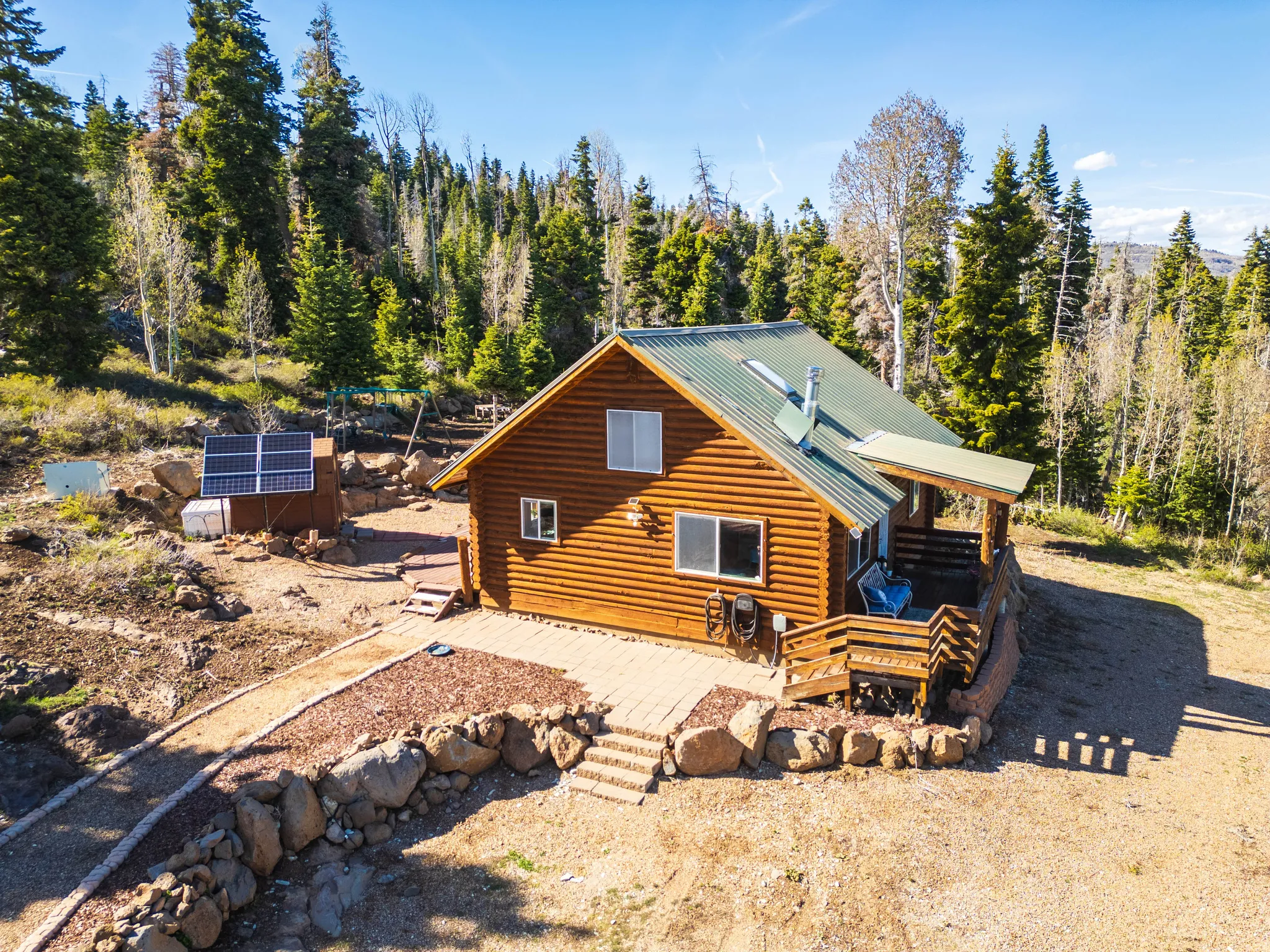 Sun-Drenched Deck & Grounds: Bright daytime aerial showing the expansive deck, solar setup, and beautifully tiered landscaping.