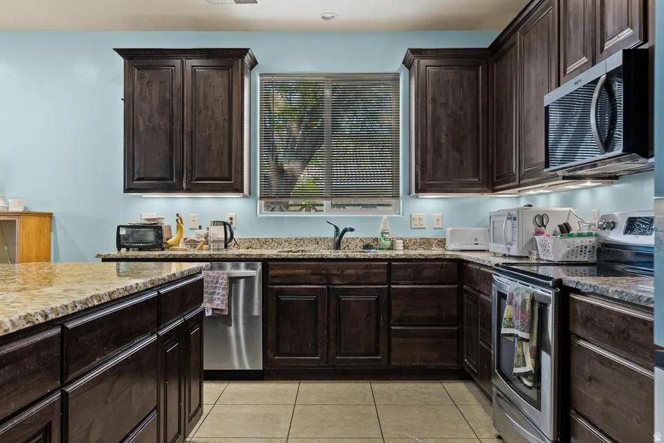 Kitchen with dark wood finish cabinets, stainless steel appliances, light tile patterned flooring, and light stone countertops