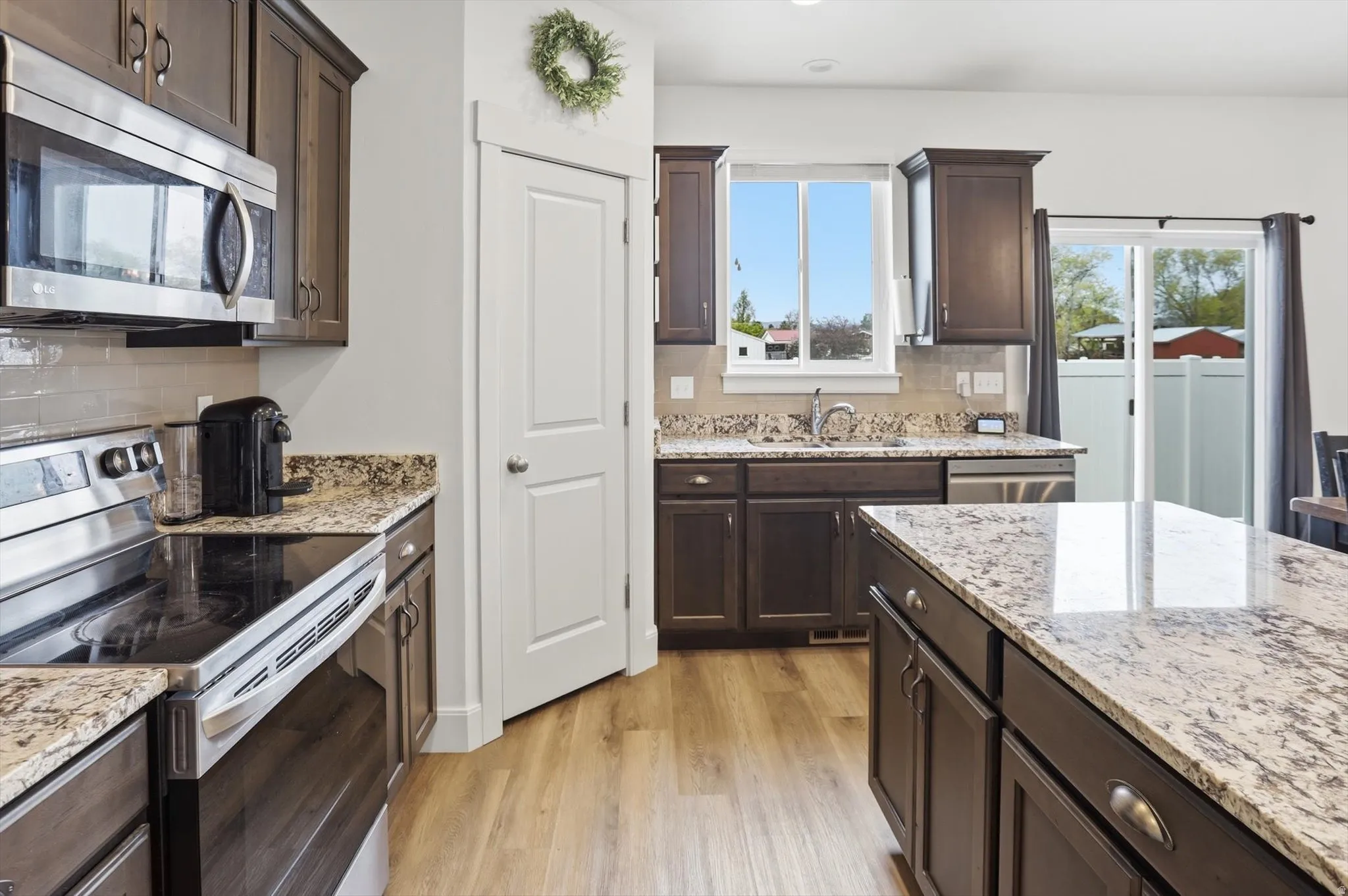 Kitchen with stainless steel appliances, dark wood finish cabinetry, light wood-style floors, light stone countertops, and backsplash