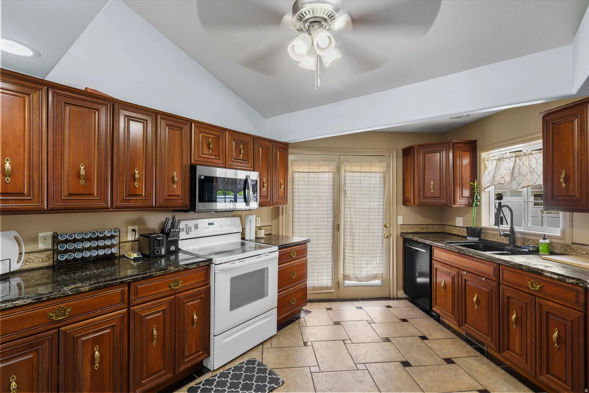 Kitchen with white electric range, vaulted ceiling, dark stone counters, stainless steel microwave, and ceiling fan