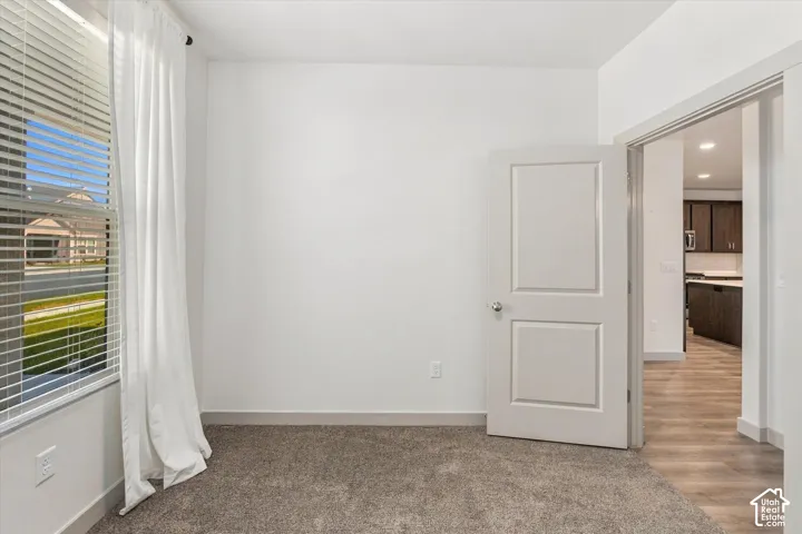 Window with blinds, wall-to-wall carpeting, white interior door, and wood-finish flooring visible in the adjacent space