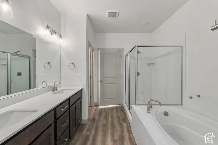 Spa-inspired primary bathroom featuring a dual-sink vanity with dark cabinetry and white quartz countertops, frameless glass shower enclosure with tile surround, freestanding soaking tub, brushed nickel fixtures, and warm wood-finish flooring throughout
