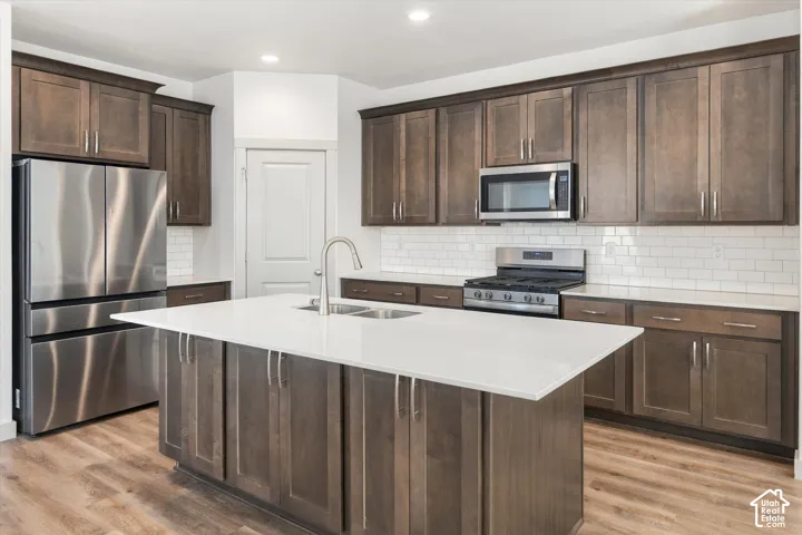 Kitchen featuring a central island with a dual-basin sink, quartz countertops, stainless steel appliances, wood-finish cabinetry, and subway tile backsplash