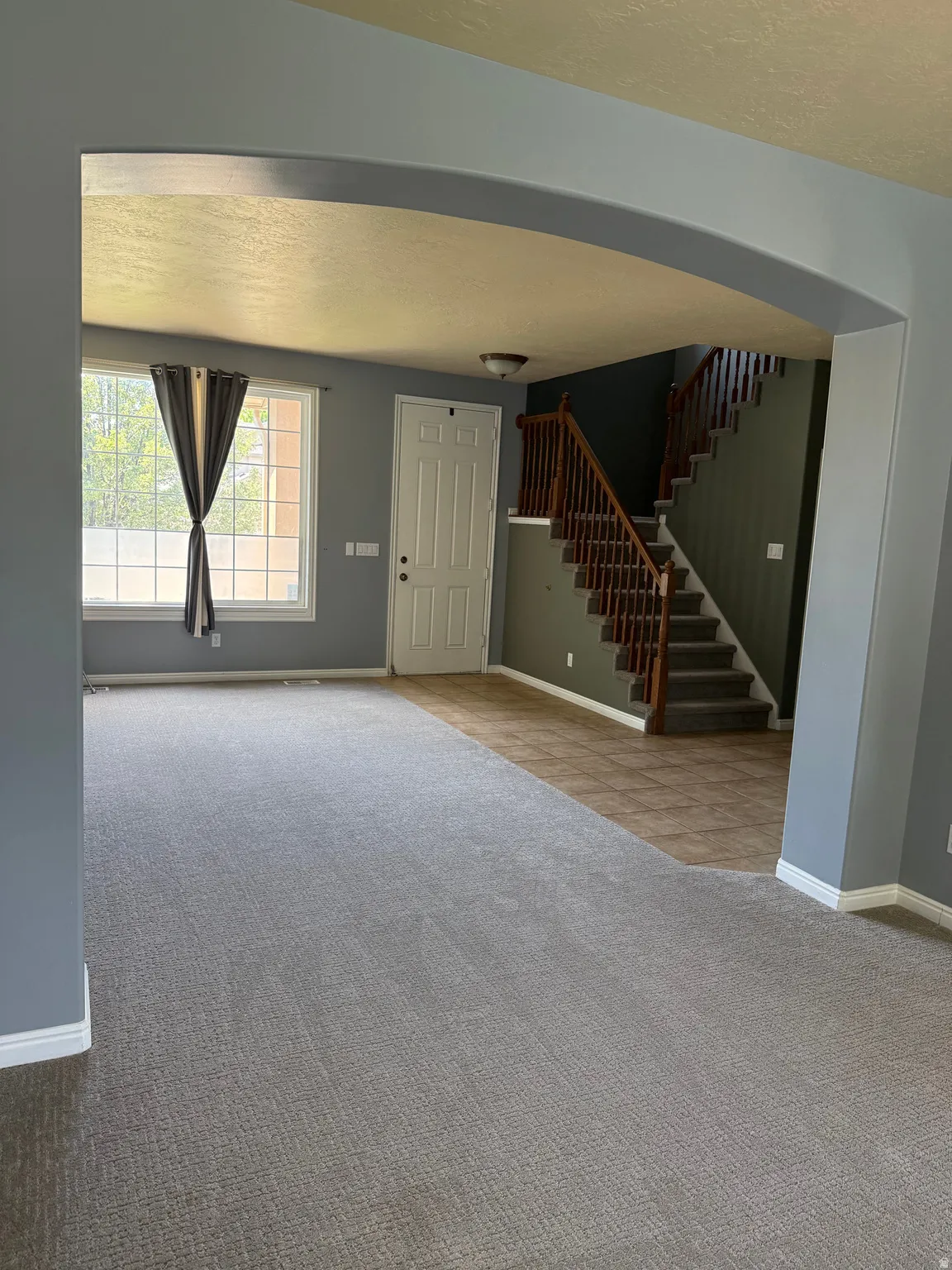 Spacious open-concept interior featuring light gray carpeting, an arched entryway, a staircase with wood railings, a white paneled front door, and a multi-pane window