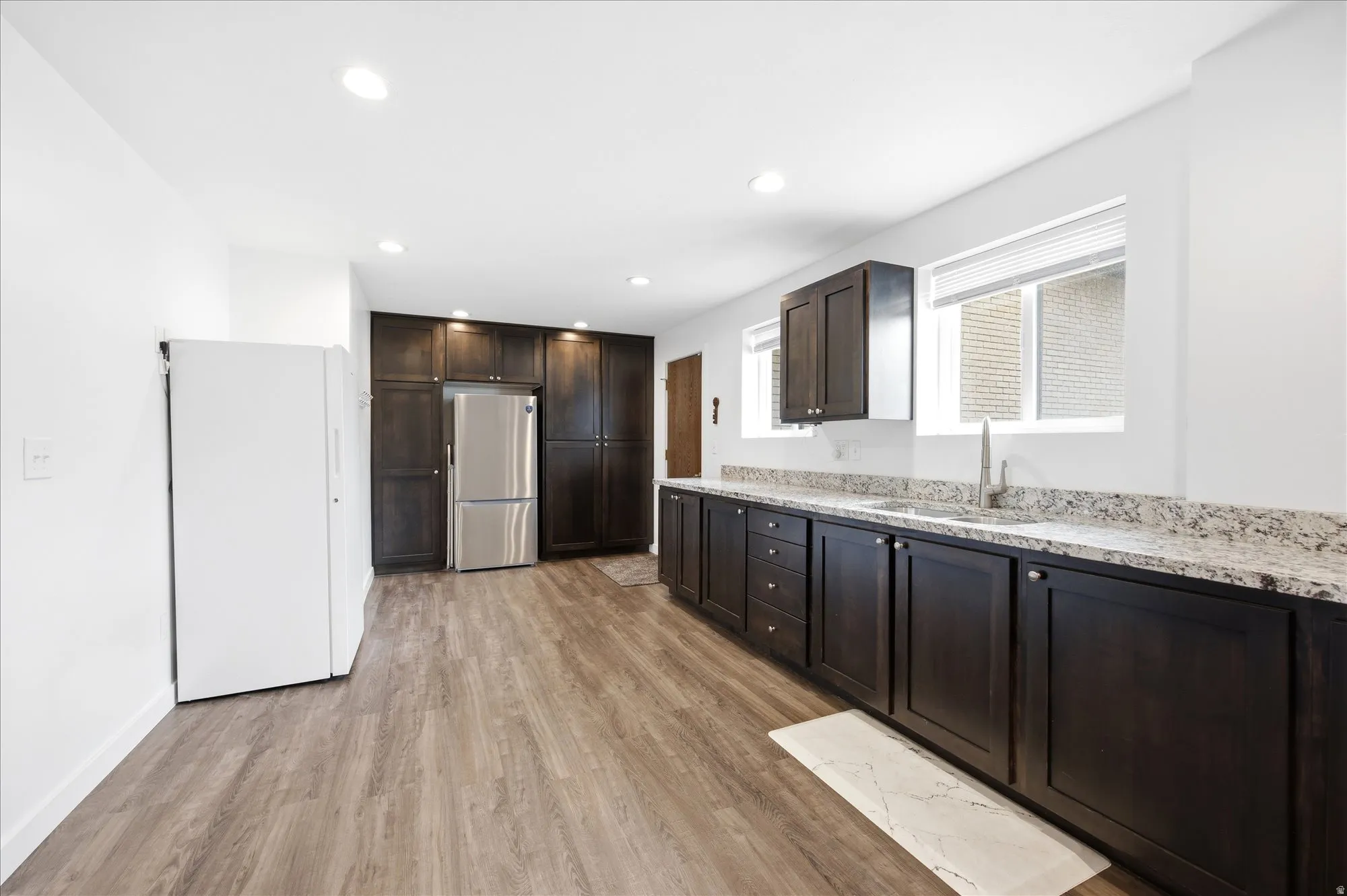 Kitchen featuring freestanding refrigerator, light wood-type flooring, dark wood finish cabinets, recessed lighting, and light stone counters
