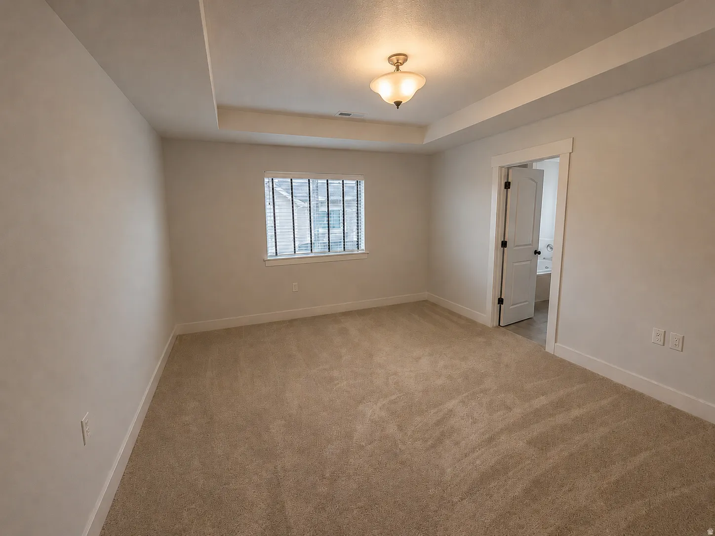 Spacious room featuring tray ceiling details, recessed lighting, neutral carpet flooring, a window with horizontal blinds, and a paneled interior door with dark hardware