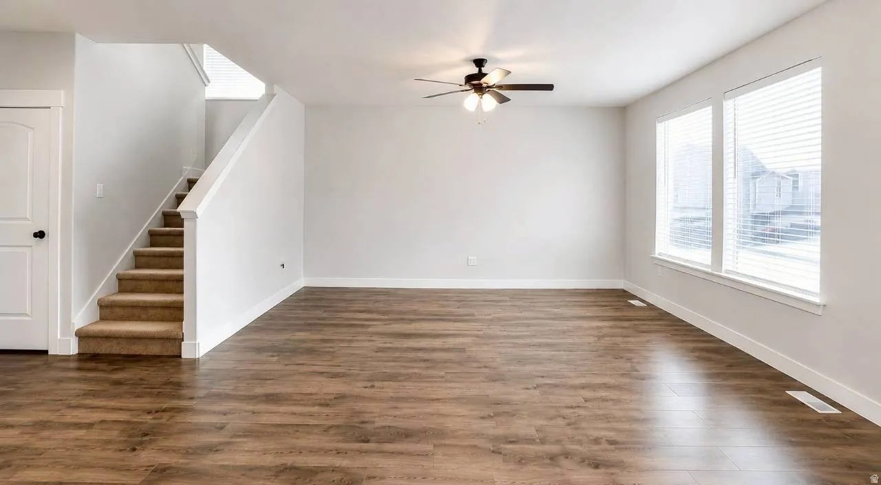 Expansive room featuring wood-finish flooring, a contemporary ceiling fan with integrated lighting, and dual large windows with blinds
