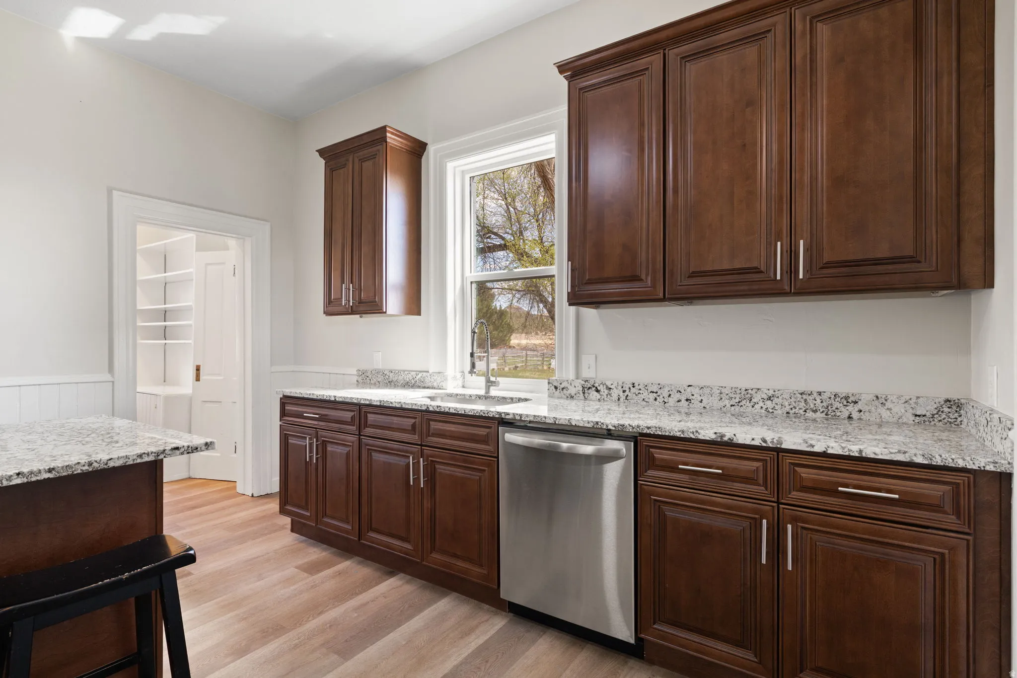 Kitchen featuring light stone countertops, stainless steel dishwasher, a wainscoted wall, light wood-style floors, and dark wood finish cabinets