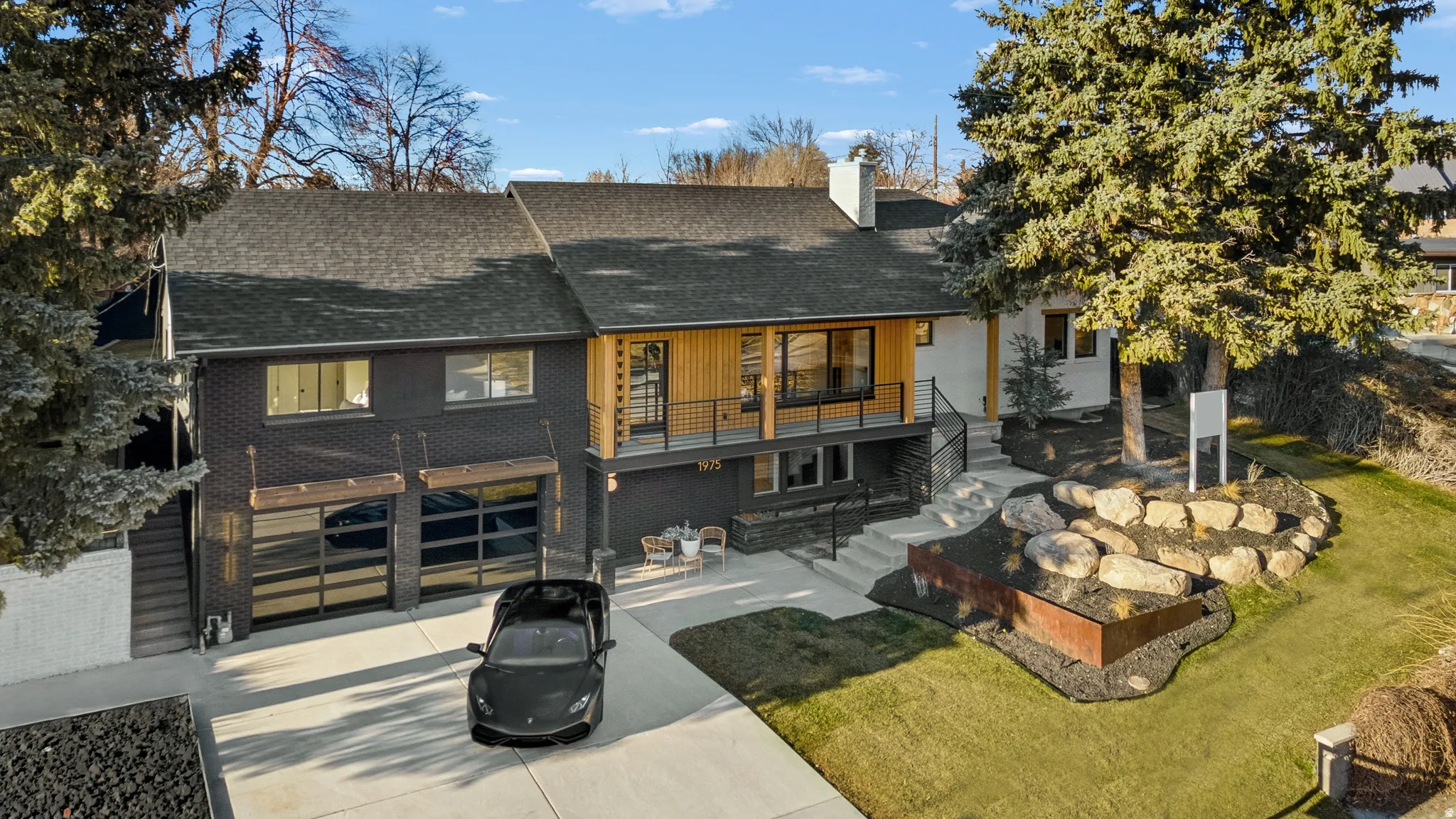 Contemporary architectural design featuring a two-car garage with glass panel doors, a prominent balcony with wood paneling, multiple exterior finishes including dark brick and light-colored siding, a stone-faced retaining wall, and a sloped, landscaped f