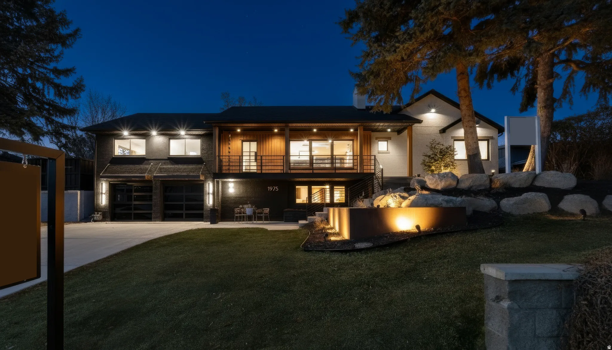 Modern exterior showcasing a two-car garage with frosted glass doors, a second-story wood-clad balcony with metal railings, and a tiered landscaped yard featuring rock elements and integrated lighting