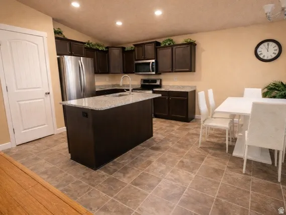 Kitchen featuring dark wood finish cabinets, stainless steel appliances, a kitchen island with sink, recessed lighting, and light stone countertops