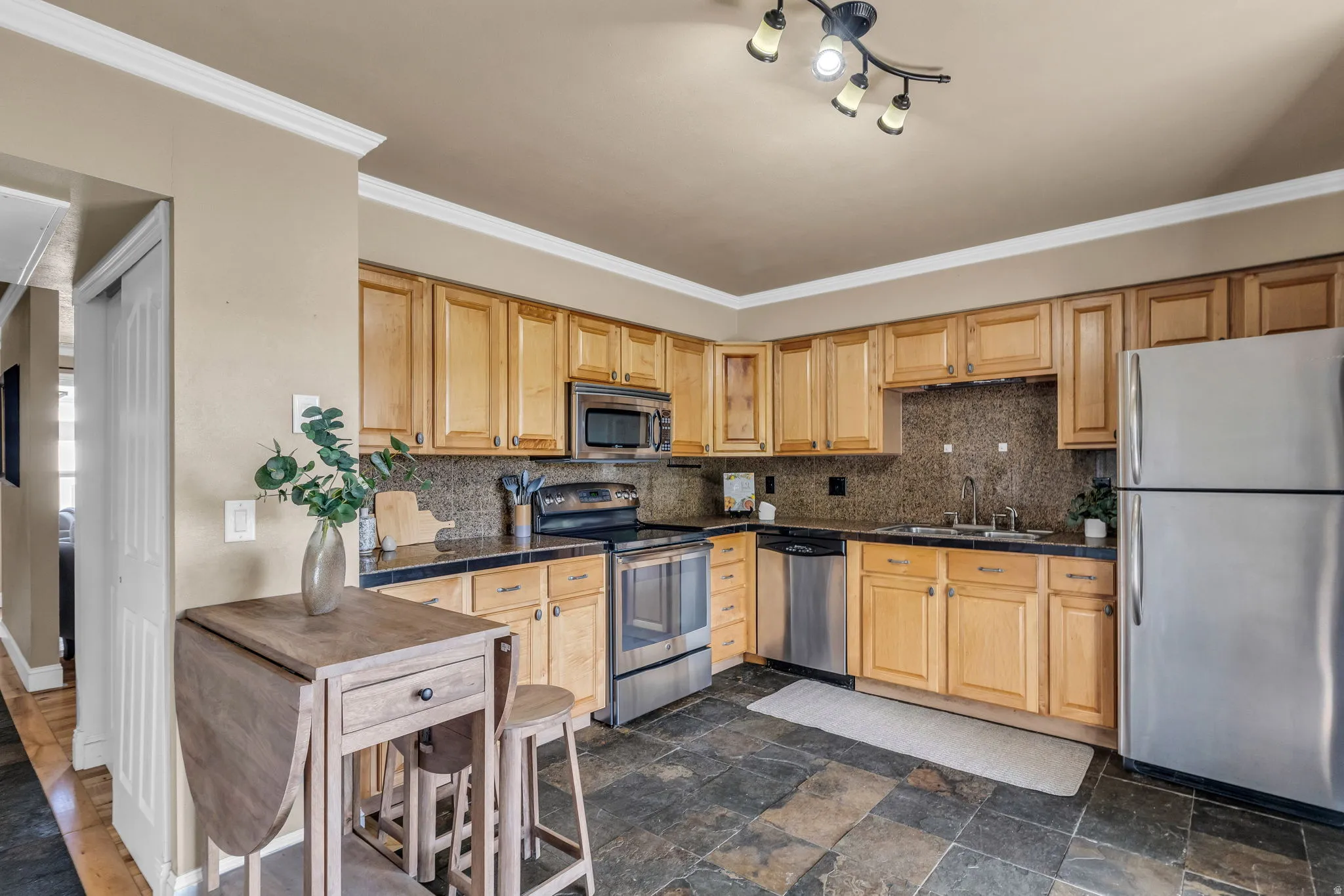 Kitchen with stainless steel appliances, light wood finish cabinetry, dark stone finish flooring, backsplash, and tile countertops