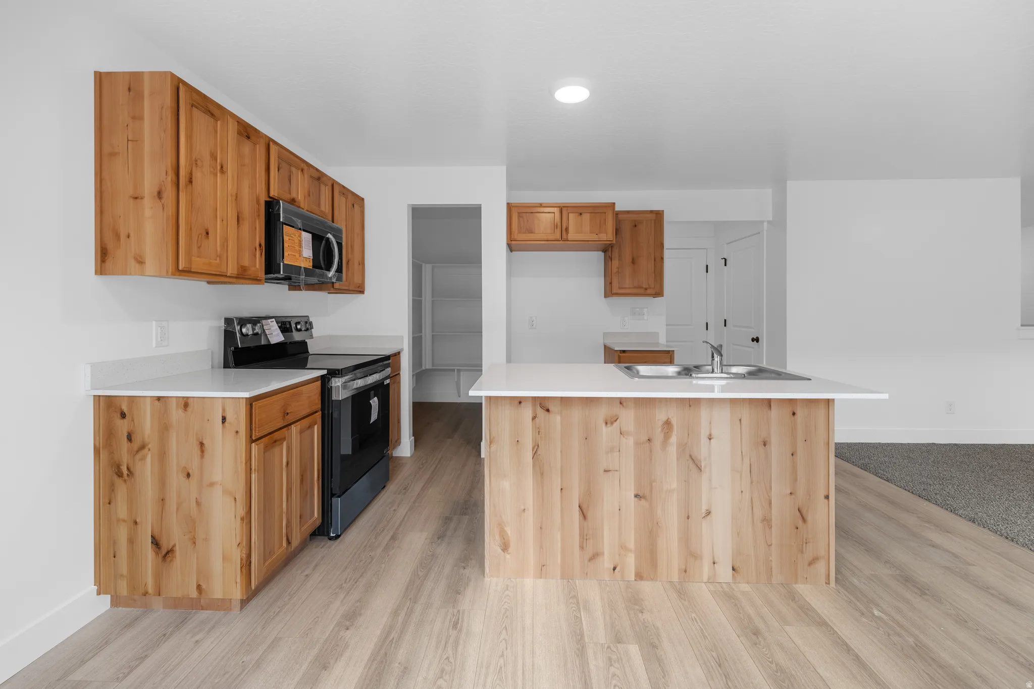 Kitchen with light countertops, stainless steel range with electric stovetop, a kitchen island with sink, black microwave, and light wood finished floors