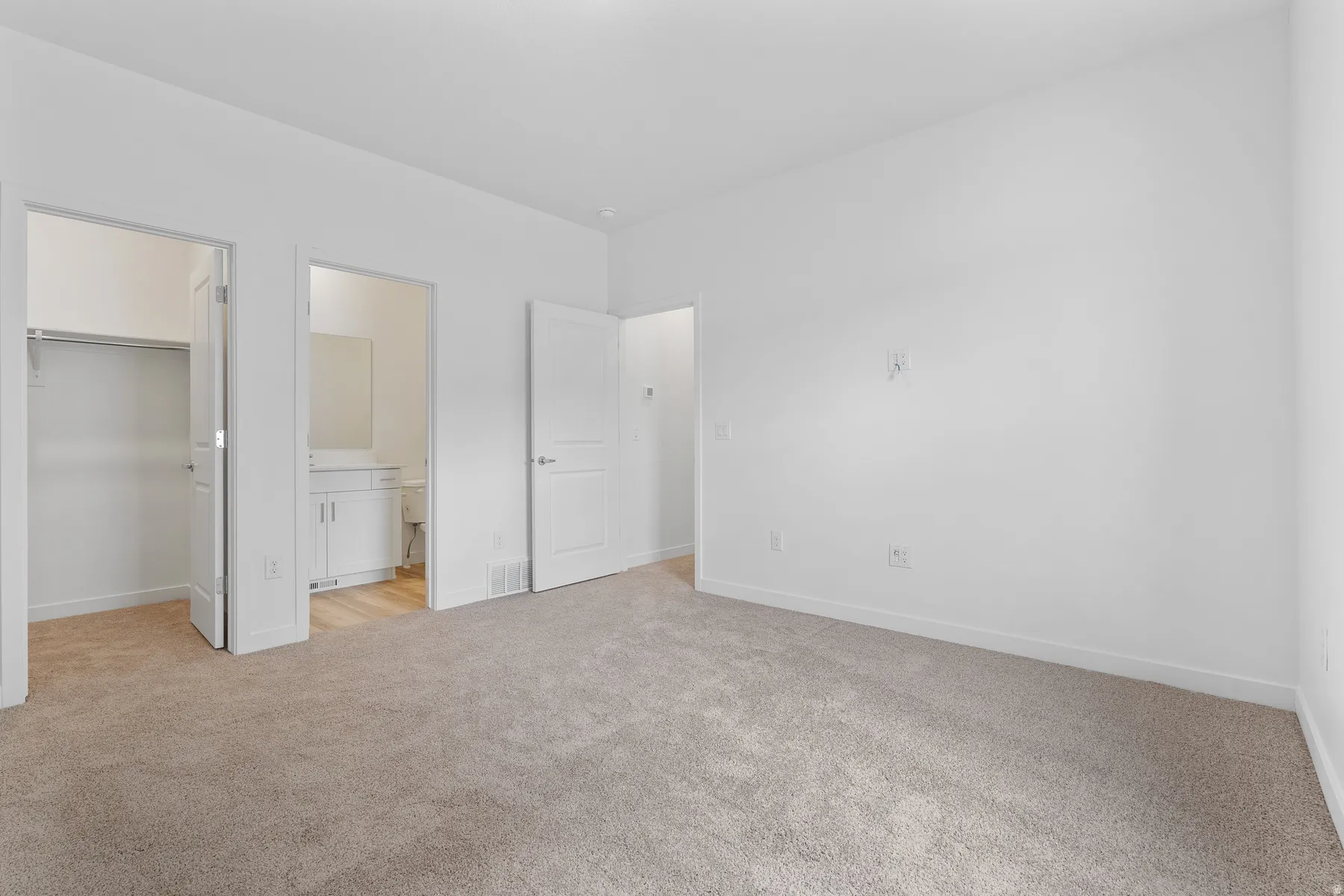 Carpeted room with white walls, featuring a built-in closet with shelving and a bathroom entry with a white vanity and mirror