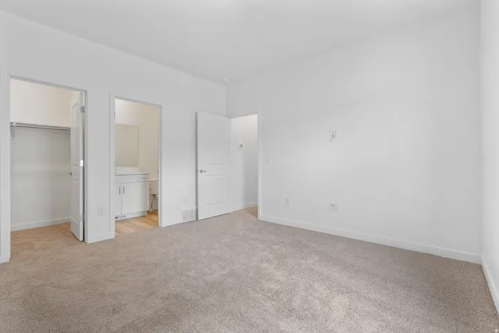 Carpeted room with white walls, featuring a built-in closet with shelving and a bathroom entry with a white vanity and mirror