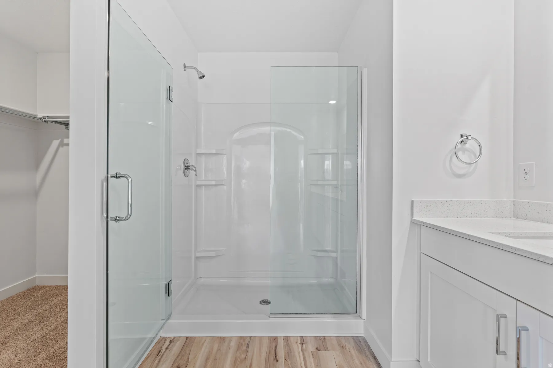 Glass-enclosed shower with white surround and integrated shelving, wood-finish flooring, and a white vanity with a light-colored countertop