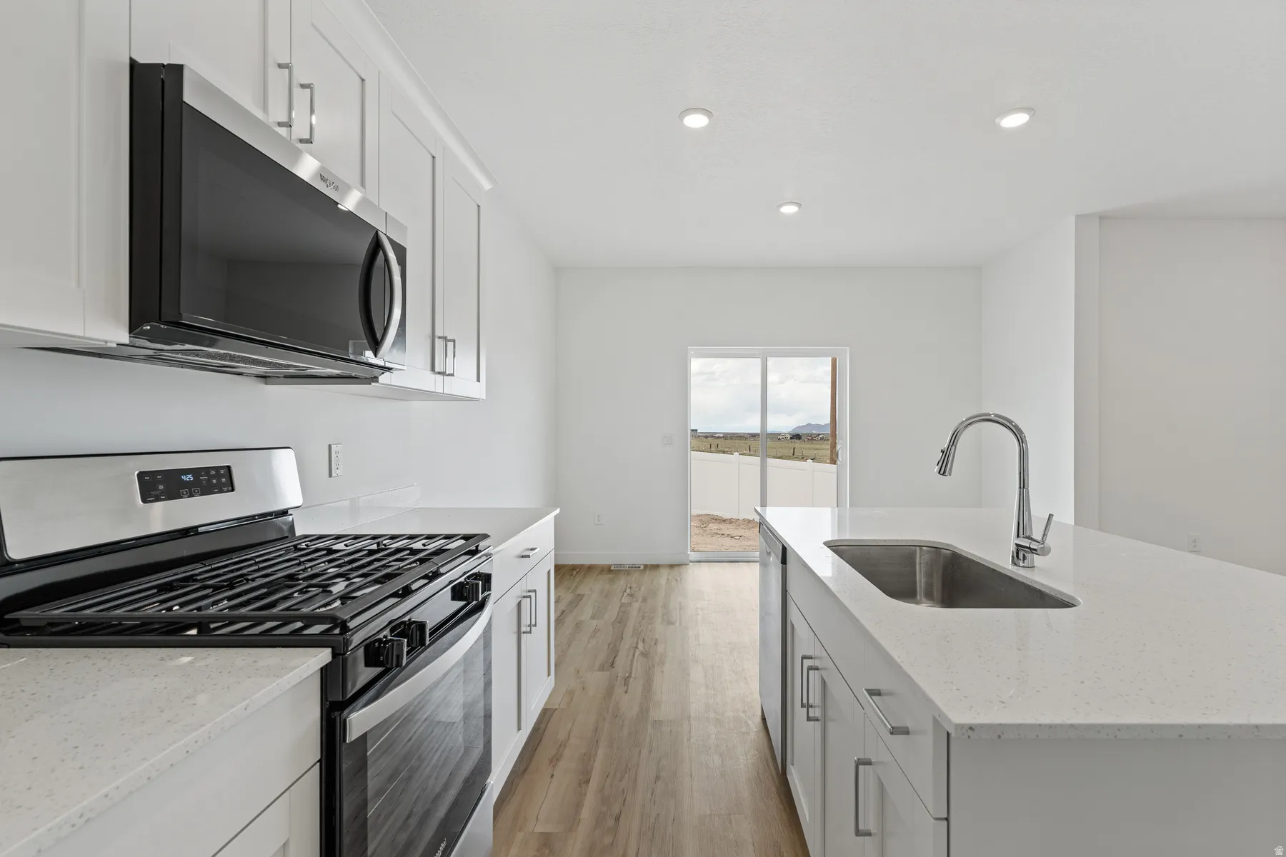 Modern kitchen featuring a stainless steel range with overhead microwave, white shaker cabinetry, light-toned countertops, and an island with an undermount sink and chrome faucet