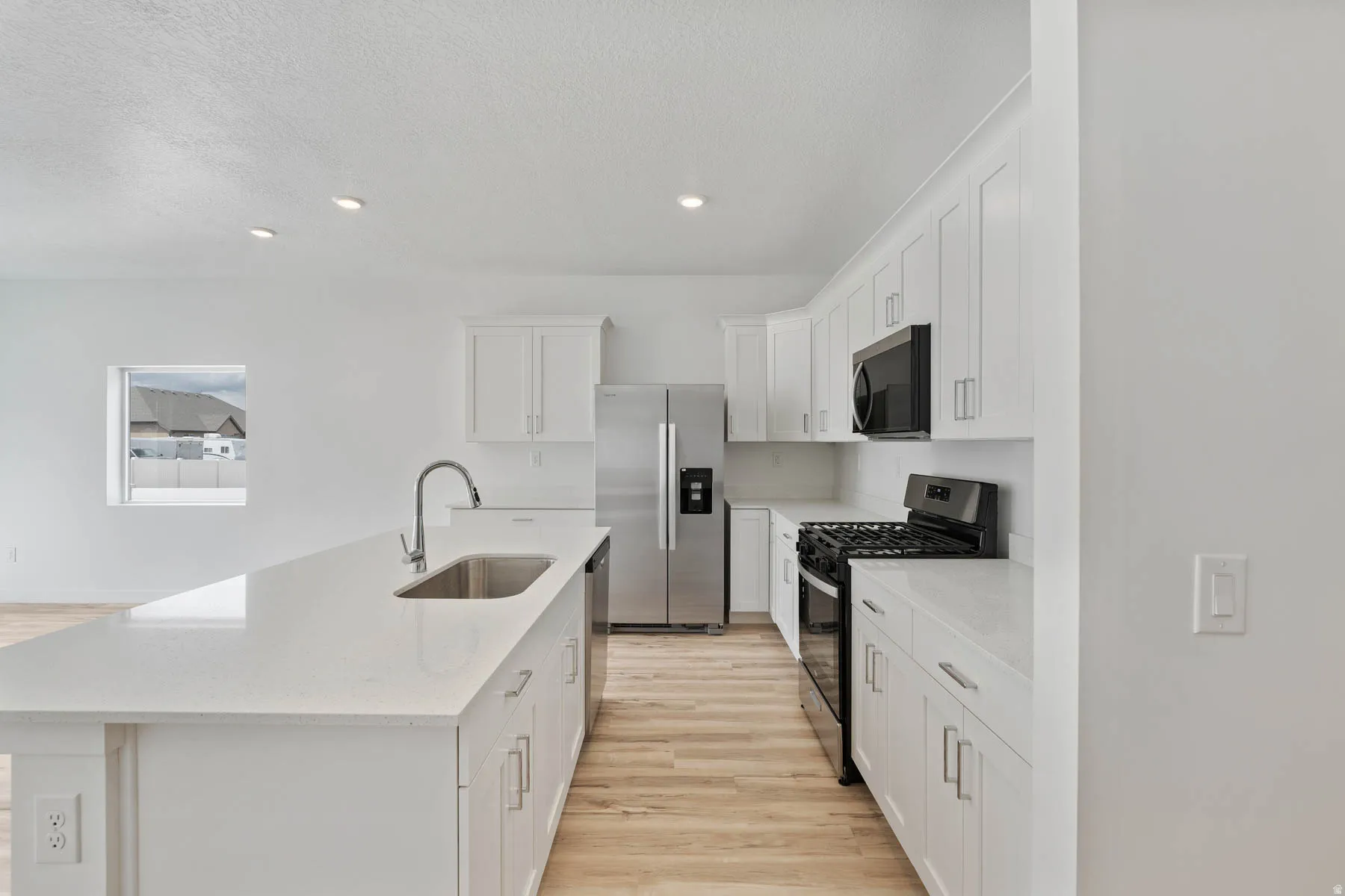 Modern kitchen featuring white shaker cabinetry, stainless steel appliances, a large center island with an undermount sink, recessed lighting, and wood-finish flooring