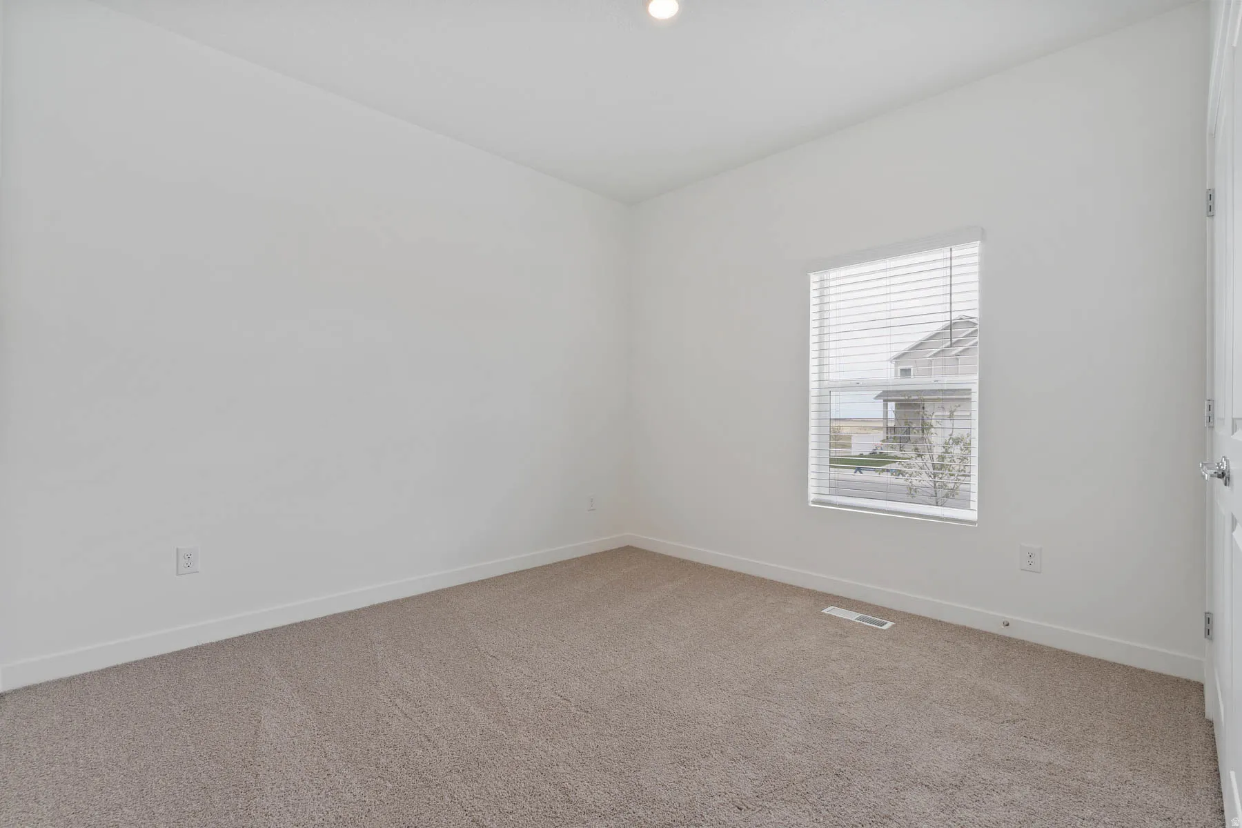 Bright interior room featuring white walls, neutral carpeting, a single window with blinds, and a white door with a silver-tone handle