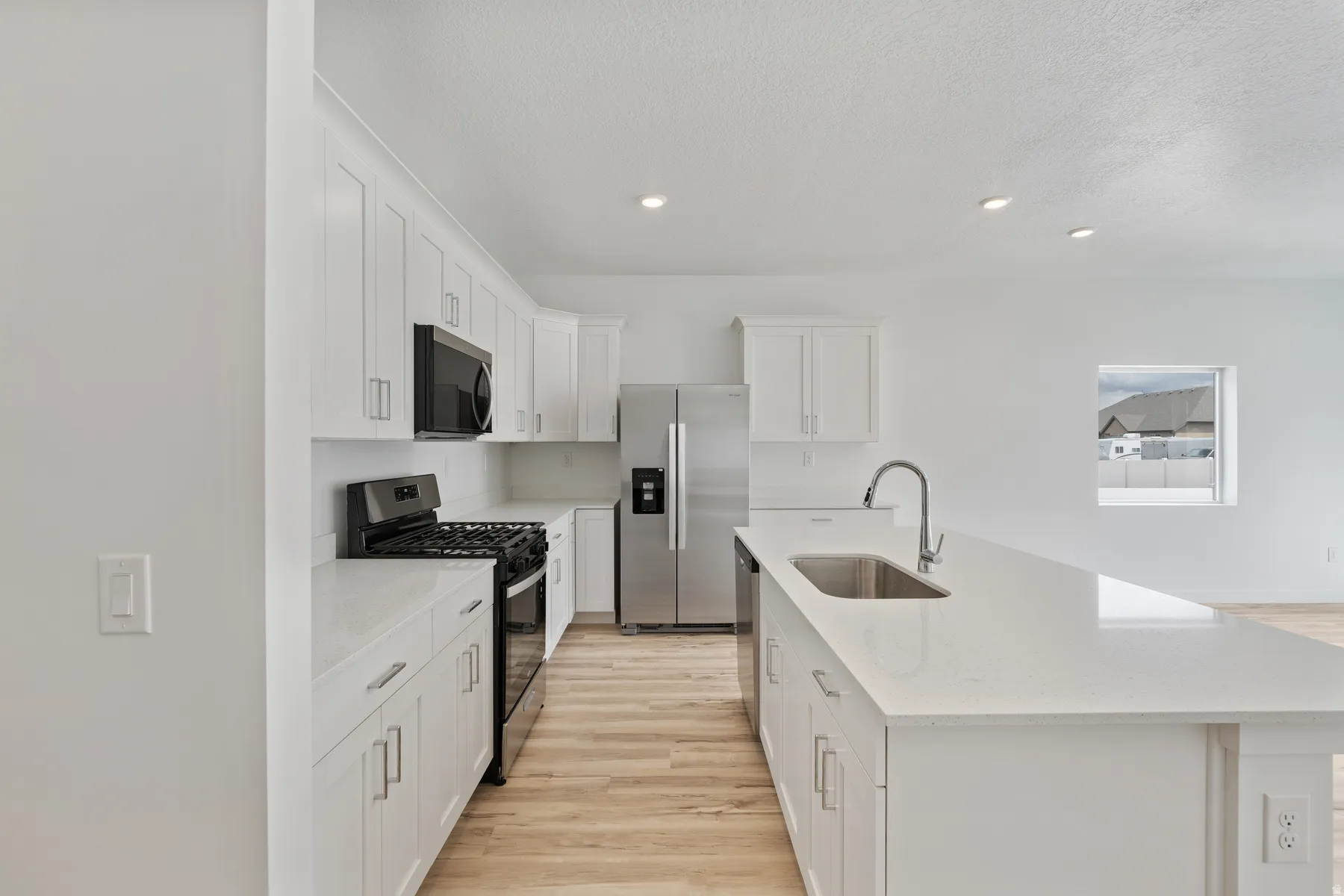 Contemporary kitchen featuring white shaker cabinetry, stainless steel appliances, light wood-finish flooring, recessed lighting, and an expansive island with an undermount sink