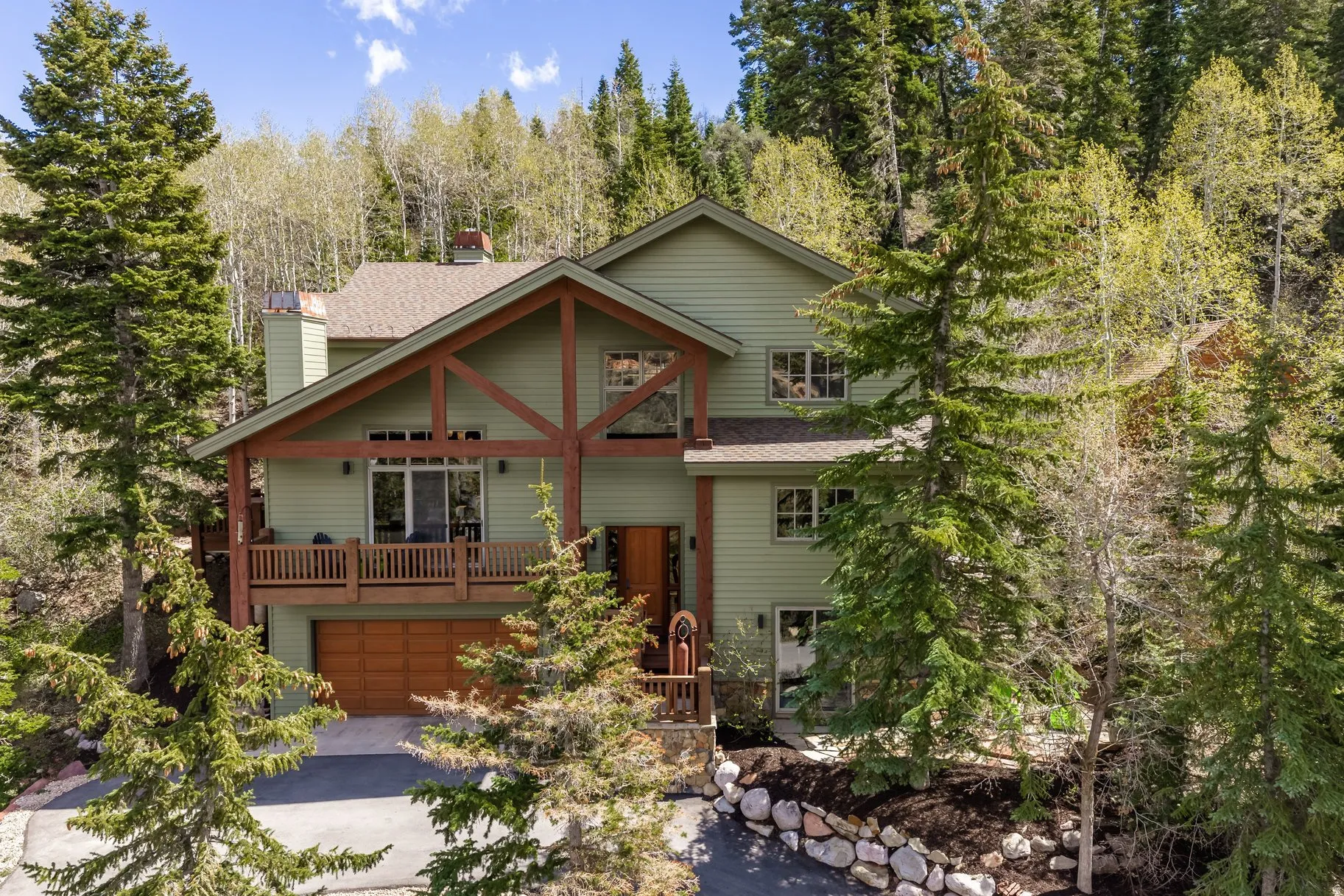 Multi-level residence featuring a sage green exterior with natural wood accents, a lower-level garage with a wood-finish door, an upper-level balcony with wood railing, and a sloped roofline