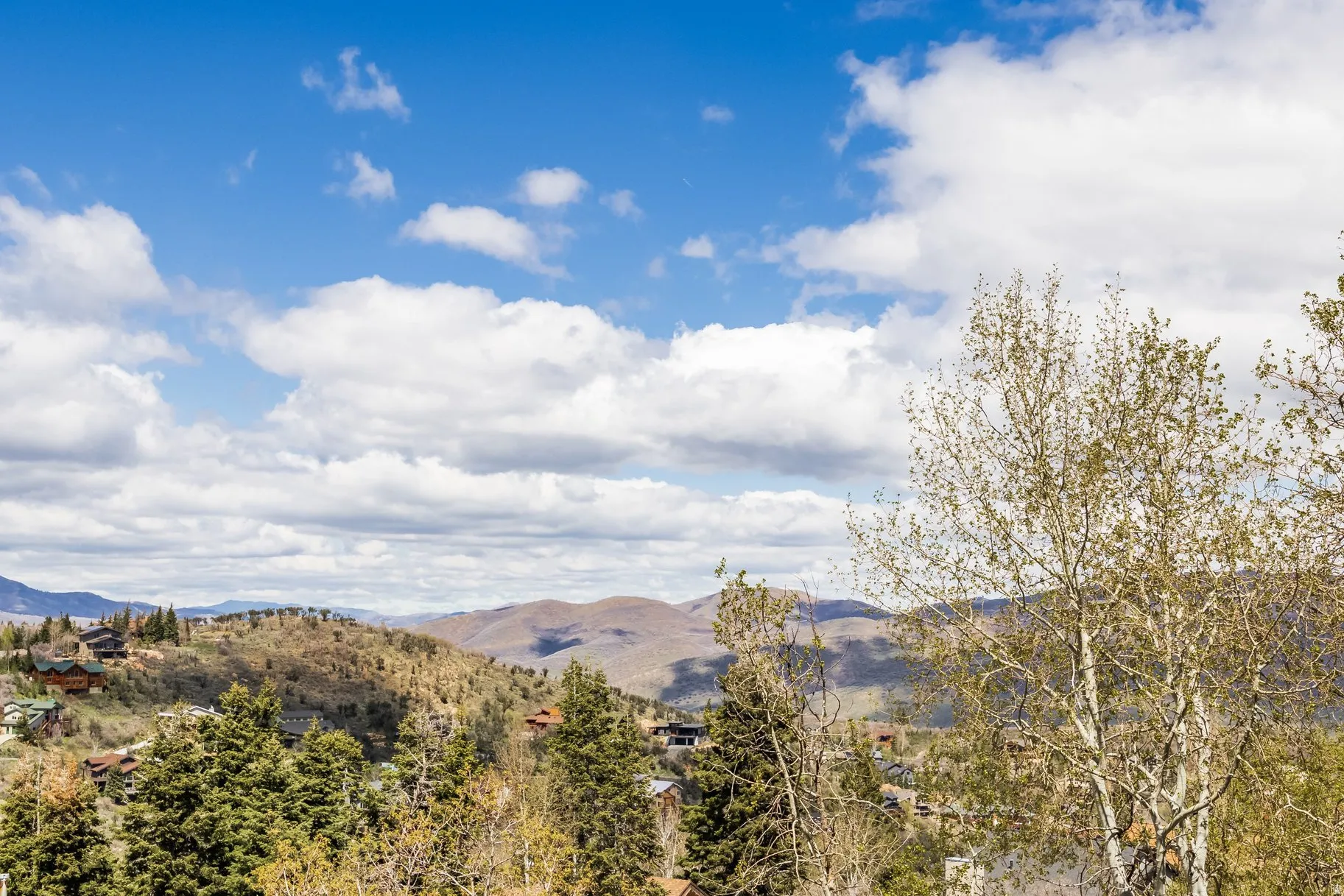 Distant mountainous terrain featuring extensive tree coverage, scattered residential structures, and a prominent blue sky with white cloud formations