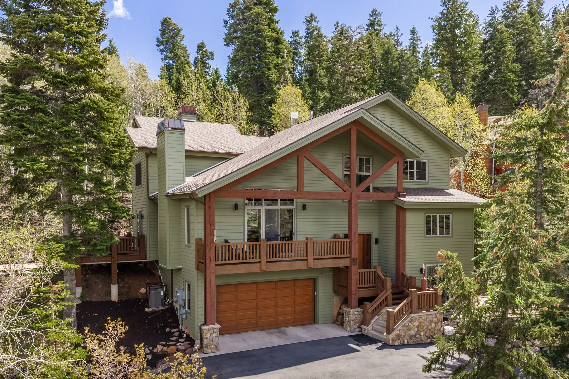 Mountain-style residence featuring a prominent timber frame entry, a full-width upper-level deck, a two-car garage with a wood-finish door, and multiple exterior staircases with stone accents