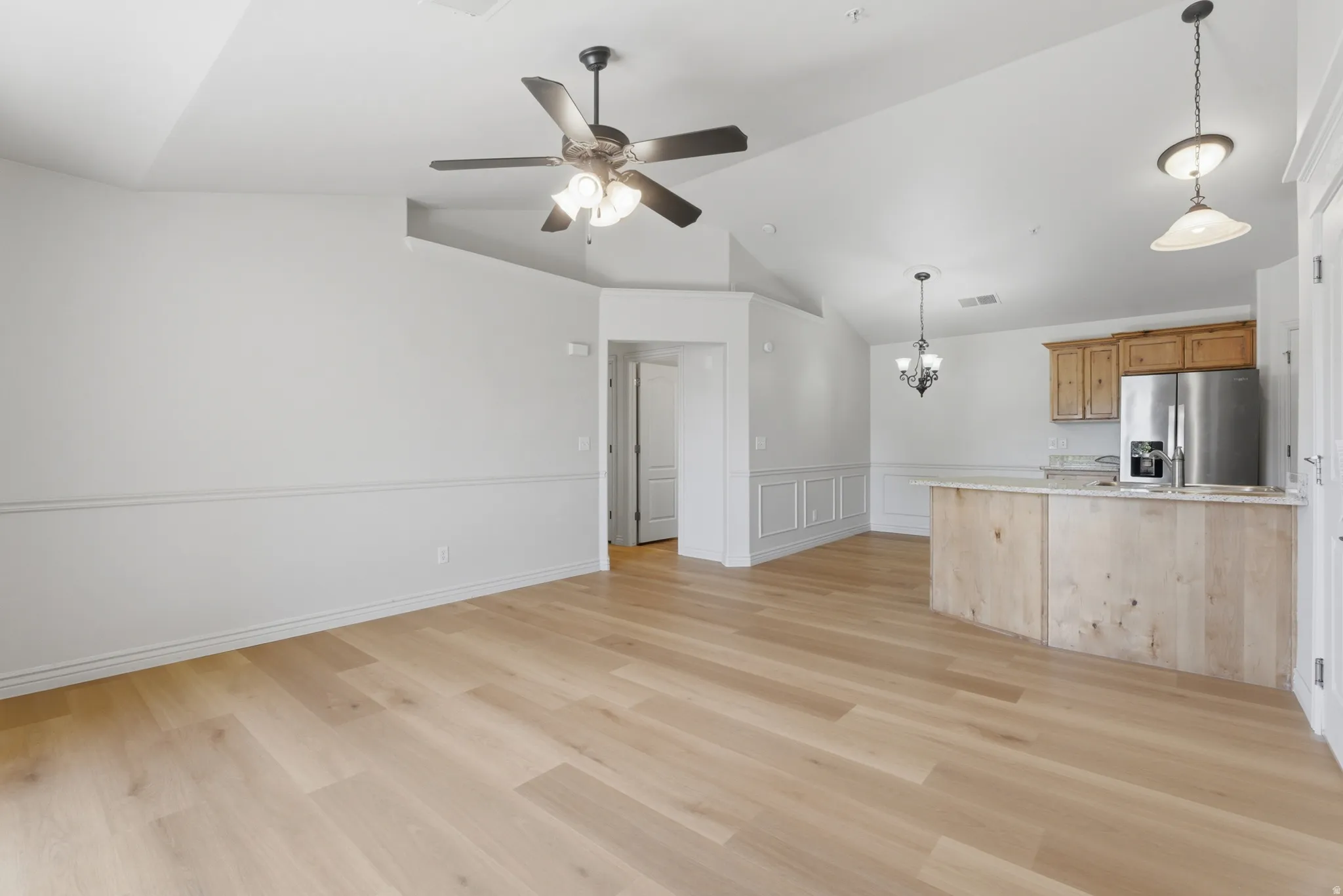 Unfurnished living room with vaulted ceiling, ceiling fan, hanging lights, light wood-style floors, and a wainscoted wall