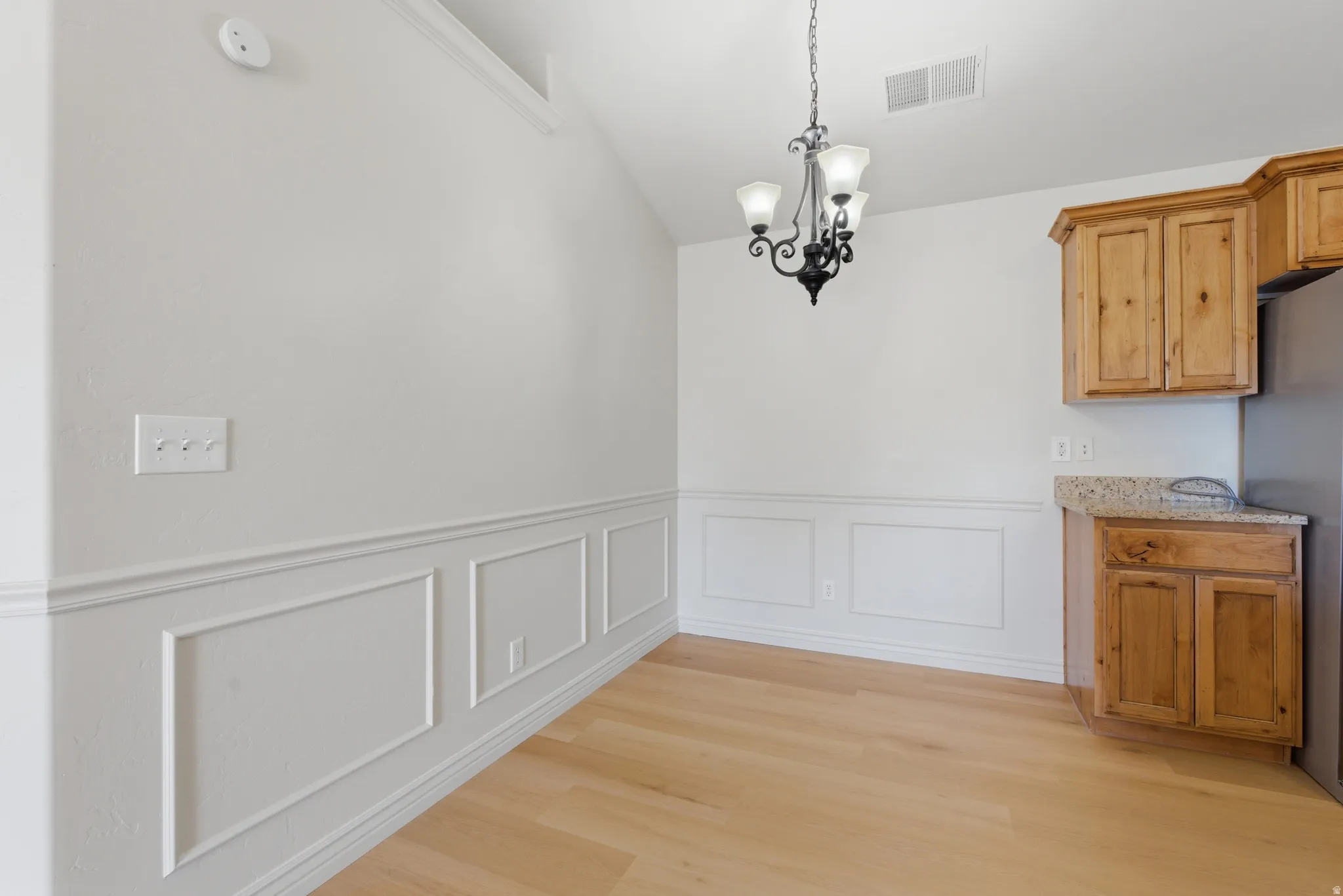 Unfurnished dining area with hanging lights, light wood-style flooring, a wainscoted wall, vaulted ceiling, and a decorative wall
