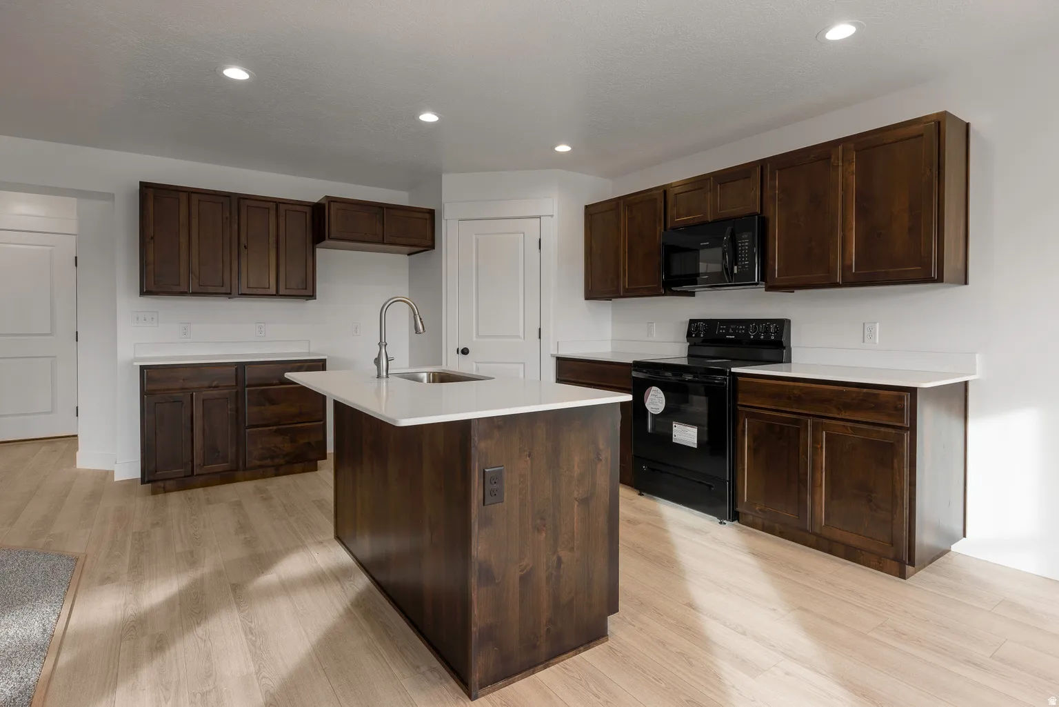 Kitchen featuring black appliances, dark wood finish cabinets, an island with sink, light wood-style floors, and recessed lighting