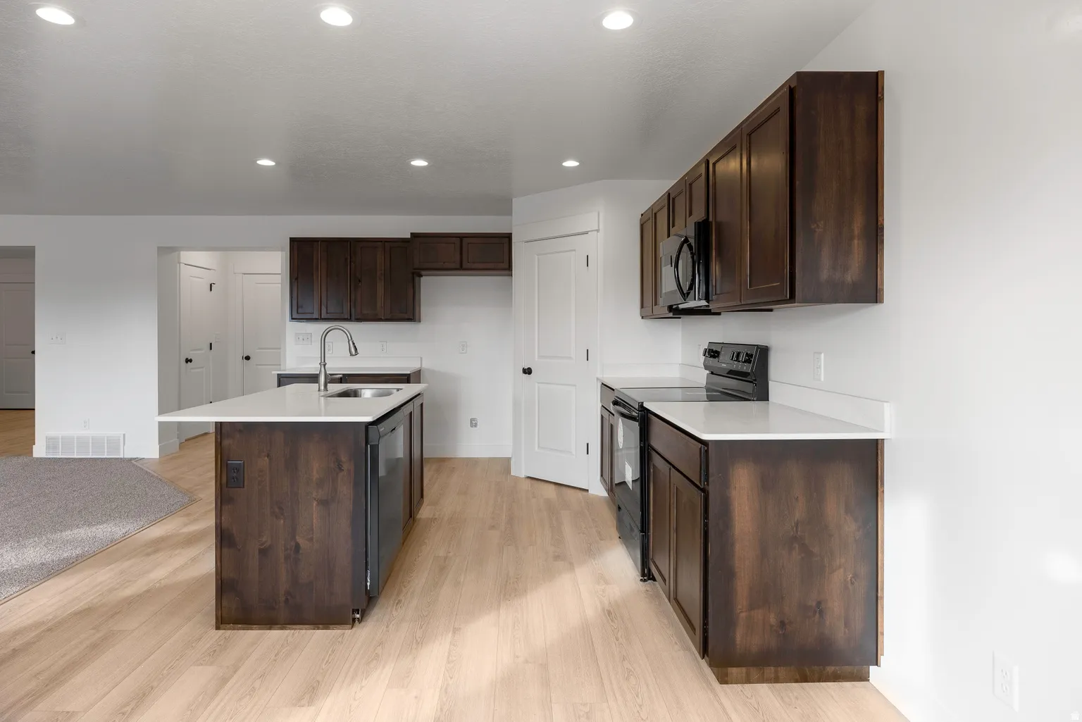 Kitchen with dark wood finish cabinets, black appliances, light wood finished floors, an island with sink, and recessed lighting