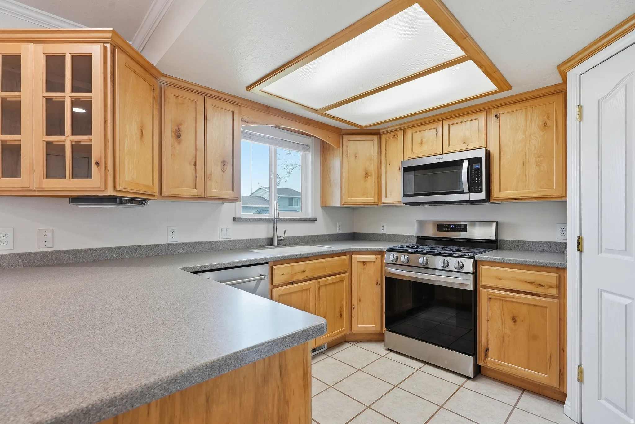 Kitchen with stainless steel appliances, glass fronted cabinets, light wood finish cabinets, light tile patterned floors, and light countertops