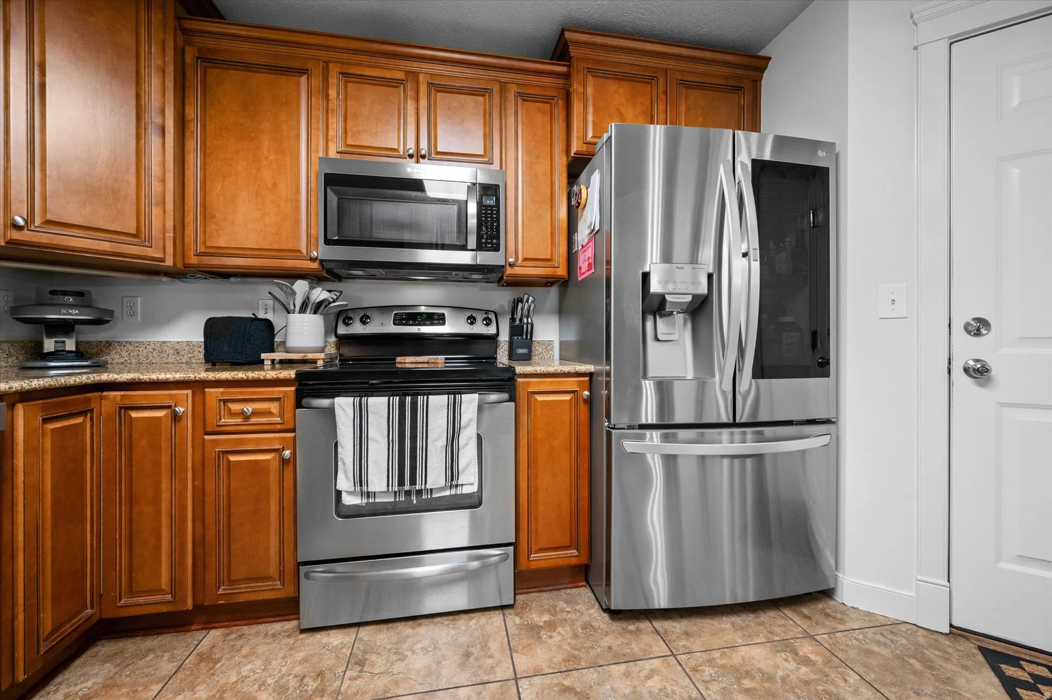 Kitchen with stainless steel appliances, wood finish cabinets, light stone counters, and light tile patterned floors