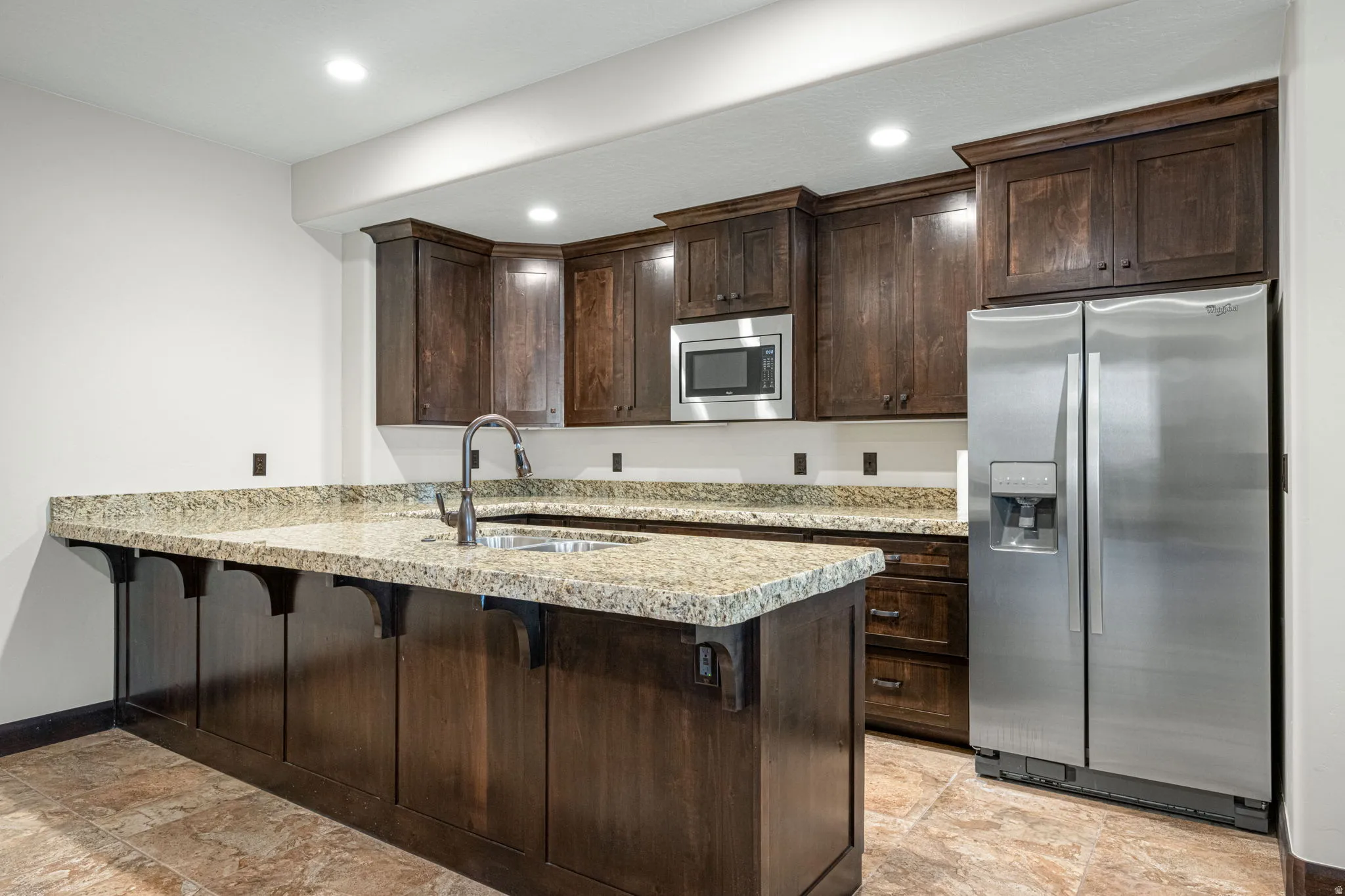 Kitchen featuring stainless steel appliances, dark wood finish cabinets, a kitchen breakfast bar, a peninsula, and light stone countertops