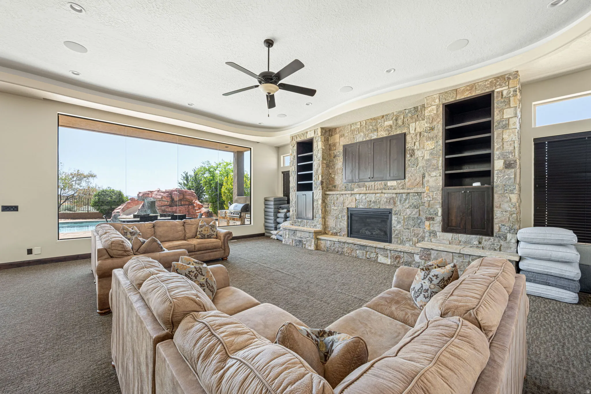 Living room with carpet, ceiling fan, built in shelves, a stone fireplace, and a tray ceiling