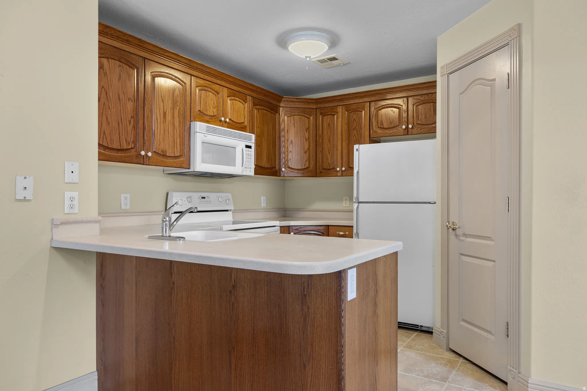 Kitchen with white appliances, wood finish cabinets, light countertops, a peninsula, and light tile patterned flooring