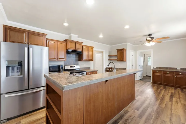 Kitchen featuring open shelves, stainless steel appliances, dark wood-style floors, crown molding, and a center island with sink